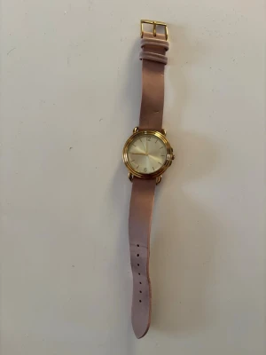 Guldig klocka med rosa läderband - Stilren klocka med rund urtavla i guld och ett rosa läderband. Urtavlan är enkel med tydliga siffror och guldfärgade detaljer. Perfekt accessoar för dig som gillar en tidlös och elegant look. Behöver nytt batteri