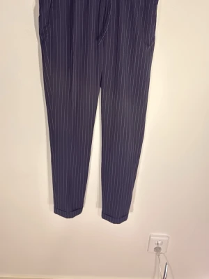 Svarta randiga kostymbyxor H&M - Säljer ett par svarta  kostymbyxor från H&M med vita tunna kritstreck. Byxorna har slim fit-passform, resår i midjan och sidofickor. Perfekta för en stilren och modern look.