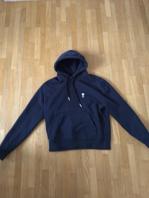 Mörkblå hoodie från Ami Paris - Mörkblå hoodie från Ami Paris med klassisk huva och dragsnören. På bröstet finns det ikoniska broderade hjärtat med bokstaven A i vitt. Hoodien har känguruficka och ribbade muddar. Perfekt för en chill och stilren look.