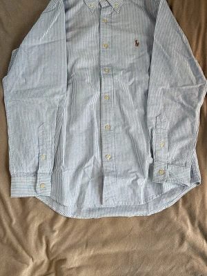 Blårandig skjorta från Polo Ralph Lauren - Säljer en klassisk blårandig skjorta från Polo Ralph Lauren i modellen Classic Fit. Skjortan har button-down krage, långa ärmar och små vita knappar. Tillverkad i bomull och har den ikoniska broderade loggan på bröstet.