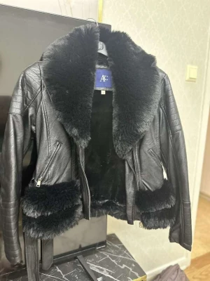 Svart skinnjacka med fuskpäls AC - Snygg svart skinnjacka från AC med bred krage och detaljer i mjuk fuskpäls. Jackan har dragkedjor på sidorna och bälte i midjan för extra stil. Perfekt för dig som vill ha en edgy look med lyxig känsla.jackan är helt ny och oanvänd.passar s och m