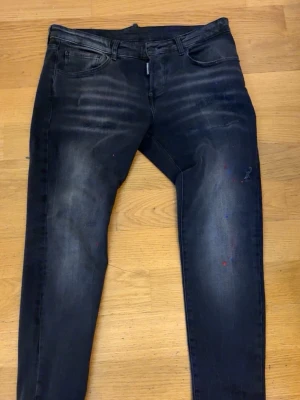 Svarta slim fit jeans från Dsquared2 - Snygga svarta slim fit jeans från Dsquared2 med slitningar och diskreta färgstänk. Jeansen har klassisk femficksdesign, dragkedjegylf och tydliga tvättade detaljer framtill. Perfekta för en trendig streetwear-look.