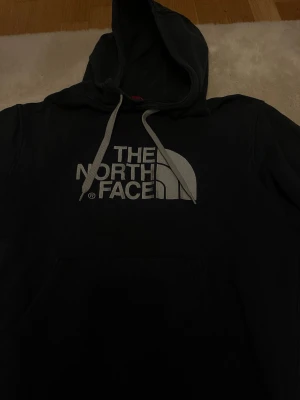 Svart hoodie från The North Face - Svart hoodie från The North Face med stor vit logga på bröstet och justerbara snören i huvan. Klassisk känguruficka framtill och mjukt material som känns skönt mot huden. Perfekt för en avslappnad och cool stil.