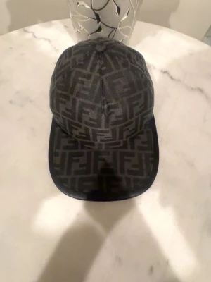 Svart keps med Fendi-logga - Snygg svart keps från Fendi med det klassiska FF-monogrammet över hela kepsen. Kepsen har böjd skärm och är tillverkad i ett slitstarkt tygmaterial. Perfekt accessoar för dig som vill ha en exklusiv och trendig look.