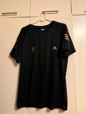 Svart Adidas t-shirt med gula detaljer - Svart sportig t-shirt från Adidas med gula detaljer och logga på bröstet samt tre gula ränder på ena ärmen. Tillverkad i funktionsmaterial (Adidas Fit System) som andas och passar perfekt till träning eller sport.