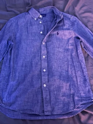 Blå linneskjorta från Polo Ralph Lauren - Säljer en snygg blå skjorta från Polo Ralph Lauren i linne med klassisk button-down krage och vita knappar. Skjortan har lång ärm och ikonisk broderad logga på bröstet. Perfekt för dig som gillar stilrena plagg med premiumkänsla i storlek 14-16 men passar mig som vanligtvis bär XS den är i väldigt fint skick ej några anmärkningar för användning .     