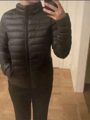 Svart dunjacka från Hollies Stockholm - Snygg svart dunjacka från Hollies Stockholm i storlek S. Jackan är lätt, packbar och har en stilren design med horisontella sömmar och hög krage. Perfekt för kalla vinterdagar och enkel att ta med sig tack vare den smidiga modellen.