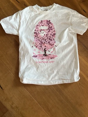 Vit BAPE t-shirt med rosa aptryck - Säljer en vit t-shirt från A Bathing Ape med ett stort rosa aptryck i form av blommor på bröstet. Klassisk passform och korta ärmar. Ikonisk BAPE-logga på etiketten vid fållen. Perfekt för dig som gillar streetwear och snygga prints.