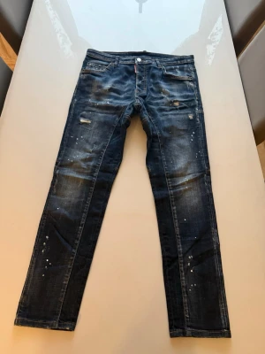Mörkblå distressed jeans Dsquared2 - Säljer ett par mörkblå jeans från Dsquared2 med slitningar, färgstänk och raka ben. Jeansen har klassisk femficksmodell, låg midja och detaljerade sömmar baktill. Perfekta för dig som gillar en edgy och trendig stil.