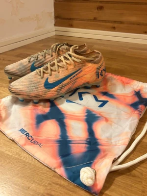 Nike Elite Kylian Mbappé fotbollsskor AG orange/blå - Säljer ett par Nike Mercurial fotbollsskor i orange och blått av Kylian Mbappé med snyggt mönster och blå swoosh (För konstgräs). Har tränat 3 gånger med dem på konstgräs och märkte att de är för stora på mina fötter. Kommer med skopåsen