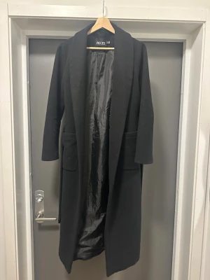 Svart lång kappa från Pieces - Stilren svart lång kappa från Pieces med klassisk krage och två stora fickor framtill. Kappan har ett bälte i midjan för snygg passform och är helfodrad på insidan. Perfekt för dig som vill ha en enkel men elegant look. Jag är 175 cm lång och den slutar under knävecket.