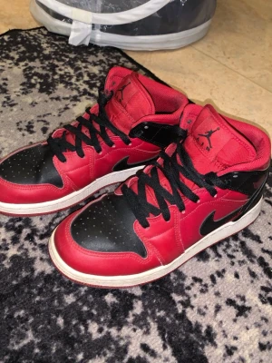 Air Jordan 1 Mid röd/svart - Snygga Air Jordan 1 Mid sneakers i rött och svart läder med svarta skosnören och vit sula. Klassisk Jumpman-logga på plösen och sidan. Skorna har en ikonisk siluett och passar perfekt till streetwear-stilen. Lite slitna o en vit fläck som inte går bort men värt priset.