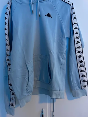 Ljusblå Kappa hoodie med logga - Snygg ljusblå hoodie från Kappa med klassiska loggor längs ärmarna och svart Kappa-märke på bröstet. Hoodien har huva med dragsko, magficka och ribbade muddar. Tillverkad i mjukt material som passar perfekt för en chill och sportig stil.