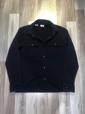Svart overshirt från Selected Homme - Stilren svart overshirt från Selected Homme med klassisk krage, knäppning framtill och två bröstfickor med lock och knapp. Tillverkad i ett mjukt tyg som passar perfekt för lager på lager. Enkel och clean design som funkar till det mesta.