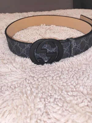 Svart Gucci bälte - Snyggt svart bälte från Gucci med klassiskt GG-mönster och stort matt svart GG-spänne. Bältet har en stilren design och är gjort i skinn med detaljer i syntet. Perfekt accessoar för att lyfta vilken outfit som helst.