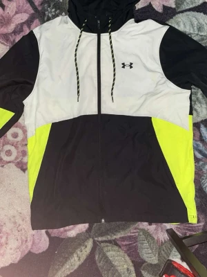 Under Armour vindjacka svart/vit/gul - Snygg vindjacka från Under Armour i svart, vitt och neongult. Jackan har huva med dragsko, hel dragkedja framtill och logga på bröstet. Tillverkad i lätt och tåligt material, perfekt för blåsiga dagar. Sportig design med färgblock och bekväm passform.