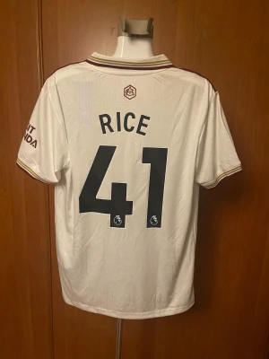 Vit Arsenal matchtröja Adidas - Säljer en vit Arsenal matchtröja från Adidas med bruna detaljer och nummer 41 samt namnet Rice på ryggen. Tröjan har klassisk krage, korta armar och klubbens logga samt sponsortryck på framsidan. Perfekt för fotbollsfans som vill ha en snygg och modern supportertröja.