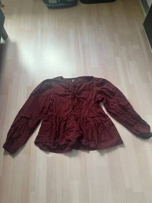 Vinröd volangblus med lång ärm - Snygg vinröd blus med volang soppskuren nederdel och långa ärmar. Blusen har en lös passform och rund halsringning. Perfekt att styla med jeans eller kjol för en trendig look.