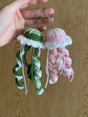 Virkat manet-nyckelring set - Två handgjorda virkade nyckelringar formade som maneter, en i grönt och vitt och en i rosa och vitt. De har spiralformade tentakler och är fästa på metallringar. Perfekt som söt accessoar till väskan eller nycklarna.