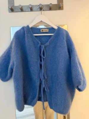 Blå fluffig kofta från Chelsea - Superfin blå kofta från Chelsea i alpacka material med knytband framtill. Koftan har korta ärmar och en rund halsringning. Perfekt för att addera lite färg till din outfit och är riktigt mysig att bära. Använd 1 gång. Nypris 990kr