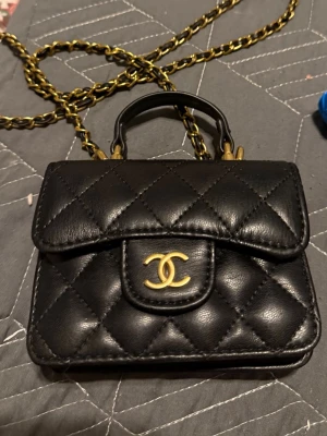 Chanel mini top handle - Säljer en svart quiltad handväska från Chanel med guldfärgade detaljer och kedjerem. Väskan har det klassiska CC-spännet framtill och är tillverkad i läder. Perfekt för dig som vill ha en ikonisk och stilren accessoar.