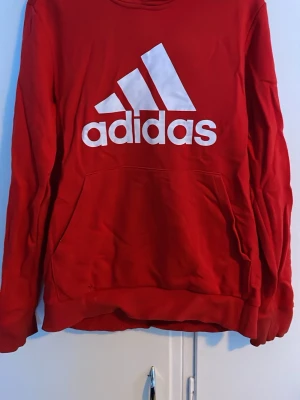 Röd hoodie från Adidas med logga - Snygg röd hoodie från Adidas med stor vit logga på bröstet. Hoodien har en klassisk känguruficka framtill och ribbade muddar vid ärmslut och nederkant. Tillverkad i mjukt material som passar perfekt för en avslappnad stil.