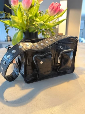 Svart axelväska med nitar NUNOO - Cool svart axelväska i croco-mönster från NUNOO med bred axelrem dekorerad med silvriga nitar. Väskan har två framfickor med dragkedja samt en stor dragkedja upptill. Perfekt för dig som gillar edgy detaljer och praktiska fack.