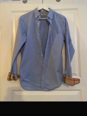 Blå skjorta från Burberry - Snygg blå skjorta från Burberry med klassisk knappkrage och bröstficka. Ärmsluten har det ikoniska rutiga Burberry-mönstret på insidan. Skjortan är i bomull och har broderad logga på bröstet. Perfekt för en stilren look.