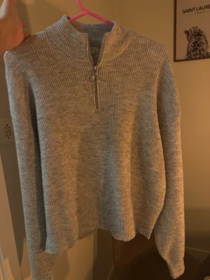 Ljusgrå ribbstickad tröja med dragkedja - Mysig ljusgrå stickad tröja med ribbad struktur och halv dragkedja framtill. Tröjan har hög krage och långa ärmar, perfekt för lager på lager. Enkel och stilren design som passar till det mesta.