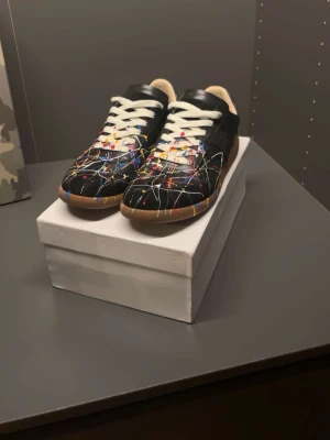 Maison Margiela sneakers med färgstänk - Unika svarta sneakers från Maison Margiela med färgstänk i flera nyanser och vita skosnören. Skorna har en klassisk låg siluett och gummisula med tydlig logotyp undertill. Perfekt för dig som vill sticka ut med din stil.
