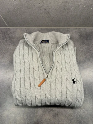  kabelstickad tröja Polo Ralph Lauren - Kabelstickad ljusgrå tröja från Polo Ralph Lauren med halv dragkedja och hög krage. Tröjan har det klassiska broderade logotypen på bröstet och ribbade muddar. Perfekt för dig som gillar stilren och tidlös design.