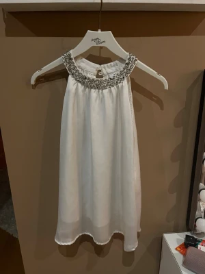 Vit ärmlös blus med pärldetaljer - Supersnygg vit blus med pärl- och paljettprydd krage. Blusen är ärmlös och har en lös, flowy passform med knapp i nacken. Perfekt för dig som vill sticka ut med en elegant touch. Materialet känns lätt och svalt.