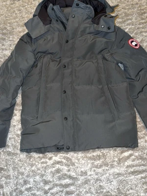 Mörkgrå dunjacka från Canada Goose - Snygg mörkgrå dunjacka från Canada Goose med klassisk patch på ärmen. Jackan har huva, hög krage och praktiska fickor framtill. Perfekt för kalla vinterdagar och håller dig varm med sin rejäla dunfyllning.