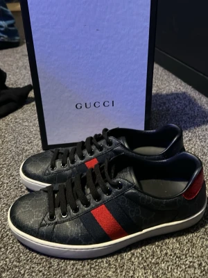 Gucci sneakers med röd/blå detalj - Snygga Gucci sneakers i mörkblått läder med klassiskt GG-mönster. Skorna har svarta skosnören, vit sula och den ikoniska röd/blå randen på sidan samt röd detalj på hälen. Perfekta för dig som vill ha en exklusiv och trendig look. Andvänt dom kanske 3 gånger inga skador förutom lite uppe på skorn som man kan se på andra bilden, pris kan diskuteras.