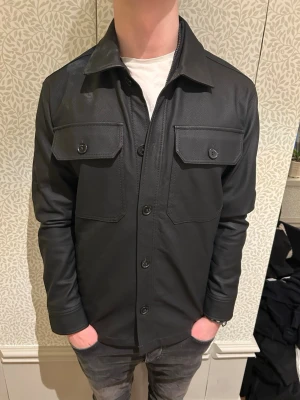 Svart overshirt i fuskläder - Säljer denna stilrena och unika Overshirt i lädermatrial perfekt inför våren. Modellen är ca 180 cm lång och väger 73 kg. Tveka inte på att kontakta mig vid minsta fundering🙌