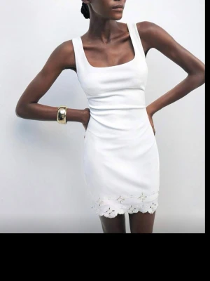 Vit kort klänning med spetskant - Säljer en söt vit klänning med en spetskant med blommönster längst ner, vilket ger en extra detalj. Aldrig använd, men säljer då den var för stor på mig. 