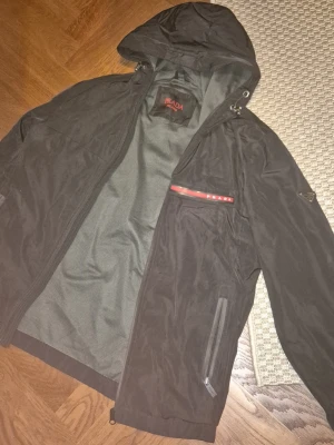Prada windbreaker - Snygg svart vindjacka från Prada med huva och dragkedja framtill. Jackan har två sidofickor med dragkedja och en bröstficka med röd detalj och Prada-logga. Tillverkad i ett lätt, vattenavvisande material och har meshfoder på insidan.