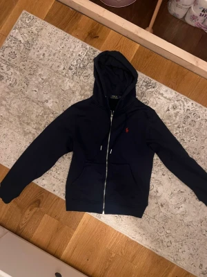 Marinblå hoodie från Polo Ralph Lauren - Snygg marinblå hoodie från Polo Ralph Lauren med dragkedja framtill och klassisk röd logga broderad på bröstet. Hoodien har huva med dragsko och två fickor framtill. Perfekt för en avslappnad och stilren look.