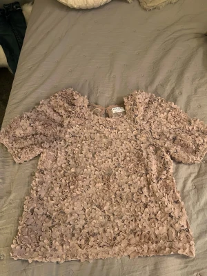 Beige blommig blus från By Malina - Supersöt beige blus från By Malina med 3D-blommor över hela tyget. Blusen har korta puffärmar och rund halsringning. Perfekt för dig som vill sticka ut med en unik och romantisk topp. Passar till både jeans och kjol!