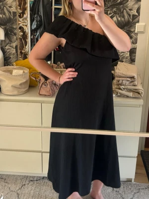 Svart oneshoulder klänning med volang - Svart oneshoulder klänning med bred volang längs ringningen. Klänningen är väldigt luftig och jag skulle säga att den passar nästan alla som har M-XL. Köpt på ullared och aldrig använd.