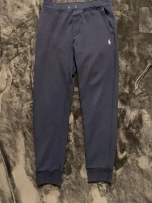 Mörkblå mjukisbyxor från Polo Ralph Lauren - Mörkblå mjukisbyxor från Polo Ralph Lauren med klassisk broderad logga på vänster ben. Byxorna har resår i midjan med dragsko, sidofickor och muddar vid bensluten. Tillverkade i mjukt bomullsmaterial för maximal komfort.