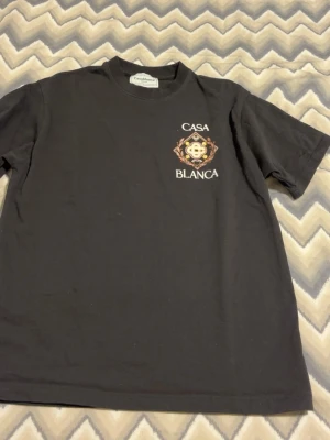 Svart Casa Blanca t-shirt med tryck - Snygg svart t-shirt från Casa Blanca med ett unikt tryck på bröstet som inkluderar logga, tennisbollar och texten 'Casa Blanca'. Klassisk passform och rund hals. Perfekt för dig som gillar streetwear och vill sticka ut lite extra.