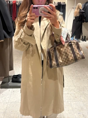 Gucci beige handväska med monogram - Säljer en beige handväska från Gucci med klassiskt GG-monogram och bruna läderdetaljer. Väskan har dubbla handtag, guldfärgade metalldetaljer och ett hänglås med nyckel. Perfekt storlek för det viktigaste och en snygg accessoar till din outfit.