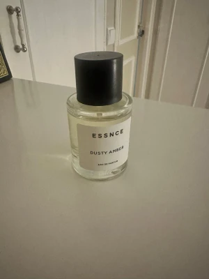 ESSNCE Dusty Amber Eau de Parfum - Minimalistisk parfymflaska från ESSNCE med doften Dusty Amber. Flaskan är genomskinlig med en enkel vit etikett och svart cylinderformad kork. Modern design som passar perfekt på parfymhyllan. Innehållet är en Eau de Parfum.