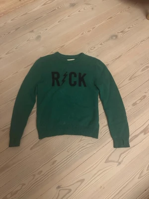 Grön cashmere tröjafrån Zadig & Voltaire med ROCK -tryck 12 år - Grön stickad tröja med svart text 'ROCK' och en blixt på bröstet. Tröjan har rund halsringning, långa ärmar och ribbade muddar vid ärmslut och nederkant. Perfekt för dig som gillar en cool och avslappnad stil. 12 år