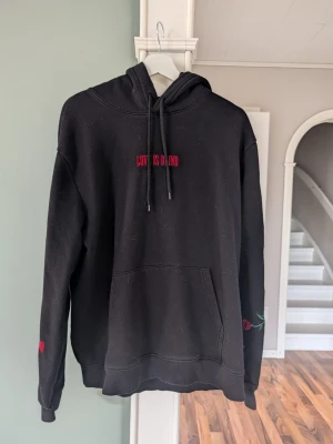 Svart hoodie med brodyr Love Is Blind - Svart hoodie med huva och dragsnören, broderad text 'LOVE IS BLIND' i rött på bröstet. Har känguruficka framtill och detaljer med broderade rosor på ena ärmen. Perfekt för dig som gillar streetwear och vill ha något unikt.