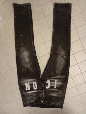 Svarta Dsquared2 ICON jeans - Säljer ett par svarta skinny jeans från Dsquared2 med vit 'ICON'-text och slitningar framtill. Jeansen har en tvättad look, detaljerade slitningar och klassiska bakfickor. Perfekta för dig som gillar streetstyle och statement-plagg. Passar 160-173
