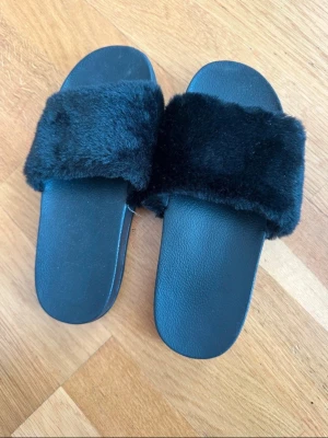 Svarta fluffiga tofflor - Säljer ett par svarta tofflor med breda, fluffiga band i fuskpäls och mjuk sula. Perfekta för att chilla hemma eller slappa runt i. Storlek EU 34/35. Klassisk slip-in modell med gummisula för extra komfort.