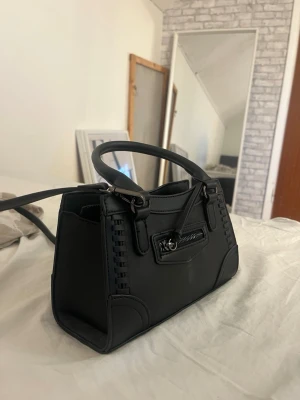 Svart handväska med dragkedjedetalj - Snygg svart handväska i konstläder med korta handtag och avtagbar axelrem. Finns inte längre att köpa på hemsidan. Väskan har en cool dragkedjedetalj framtill och dekorativa sömmar längs kanterna. Perfekt storlek för det viktigaste och ger en trendig touch till din outfit.