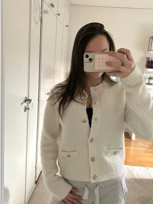 Krämvit kofta med träknappar - Mysig krämvit kofta med rund halsringning och träknappar framtill. Koftan har två små fickor med knappdetaljer och ribbade muddar vid ärmslut och nederkant. Perfekt för lager på lager och enkel att matcha till olika outfits.